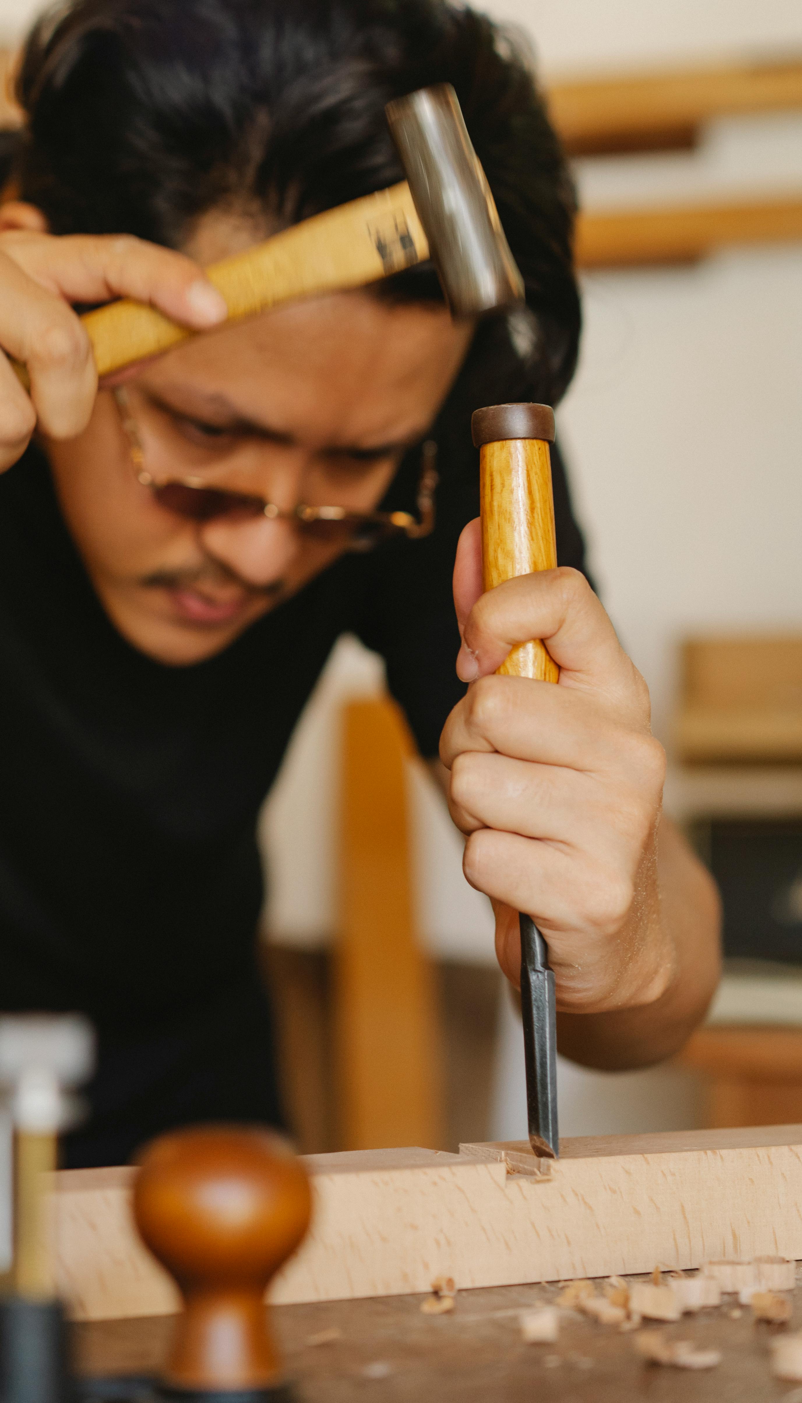 serious ethnic craftsman cutting wooden piece with chisel and hammer