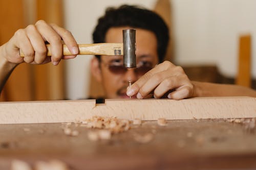 Focused ethnic male carpenter hammering nail into wooden detail in workshop
