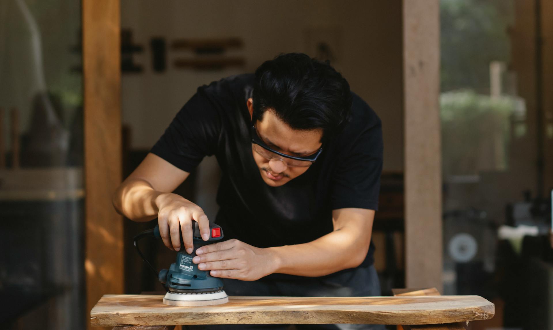 Jeune charpentier ethnique concentré en vêtements décontractés et lunettes de protection polissant des planches de bois avec une meuleuse dans l'atelier