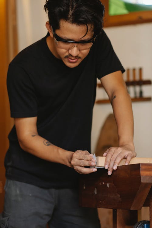 Crop young ethnic professional carpenter in casual clothes and goggles grinding edge of wooden plank in workshop