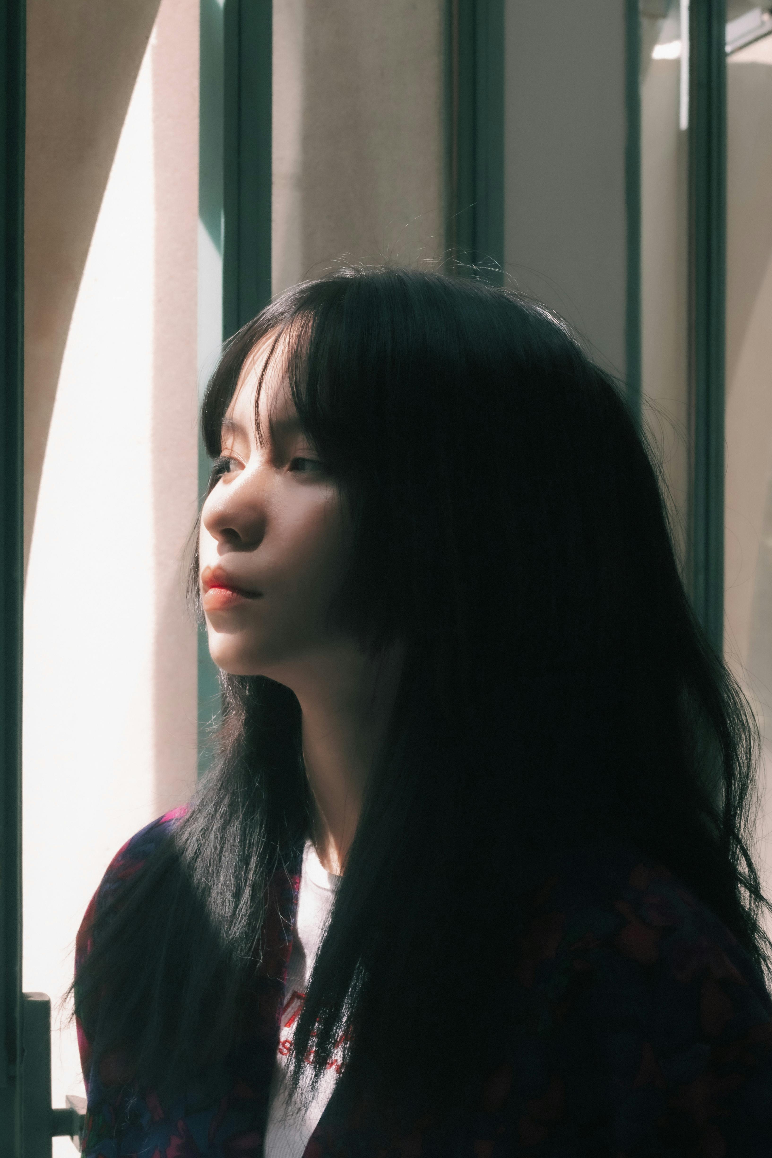 Thoughtful portrait of an East Asian woman standing by a window, bathed in gentle light.