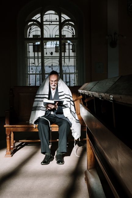 An elderly man reads a book in a synagogue, draped in a tallit, in natural light.