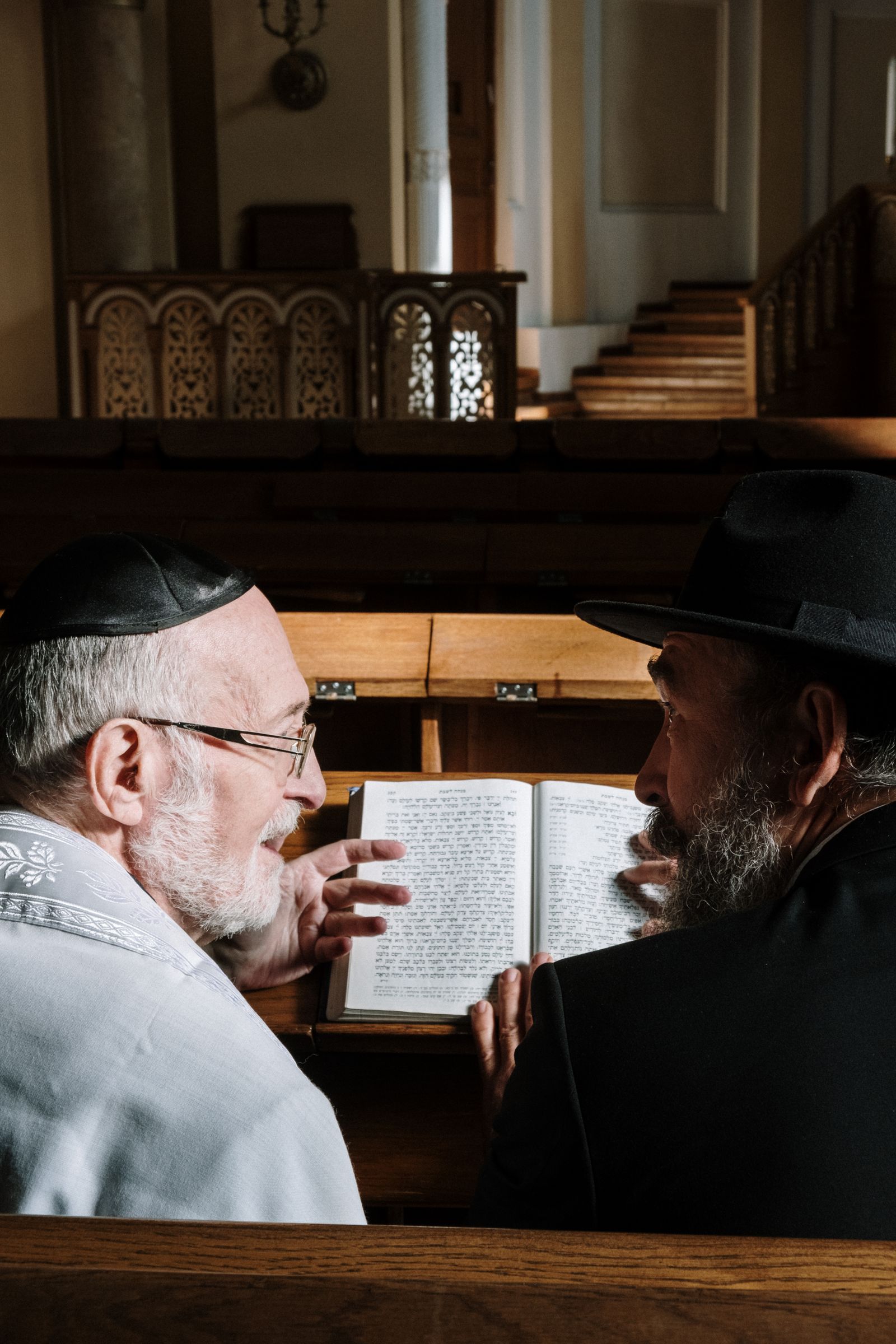 Two older men in kippot studying a book together in a synagogue pew
