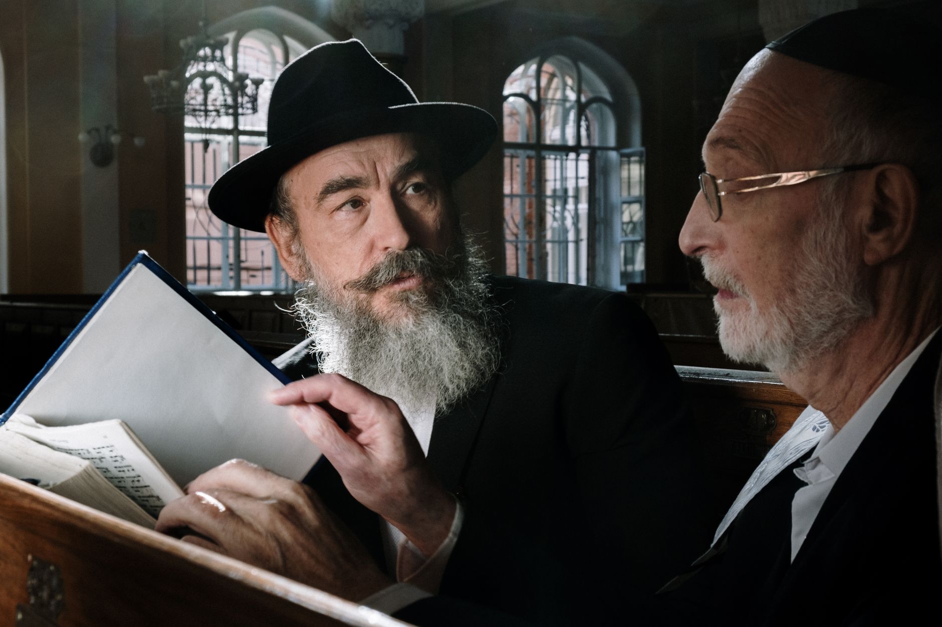 Two senior men engaging in a thoughtful conversation while reading religious texts inside a synagogue.