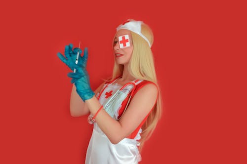 Smiling blond female nurse with ambulance cross on eye in sterile gloves with injector and stethoscope