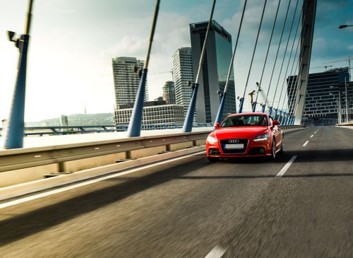 Free Vibrant red sports car driving on a modern bridge with cityscape in the background. Stock Photo