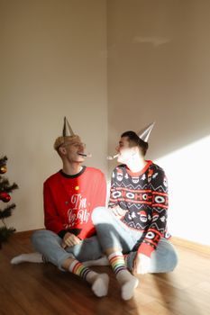两名男子身着节日圣诞毛衣，坐在地板上，用派对帽和噪音制造者在室内庆祝新年