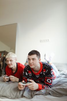 Two adults wearing Christmas sweaters playing video games on a bed indoors.
