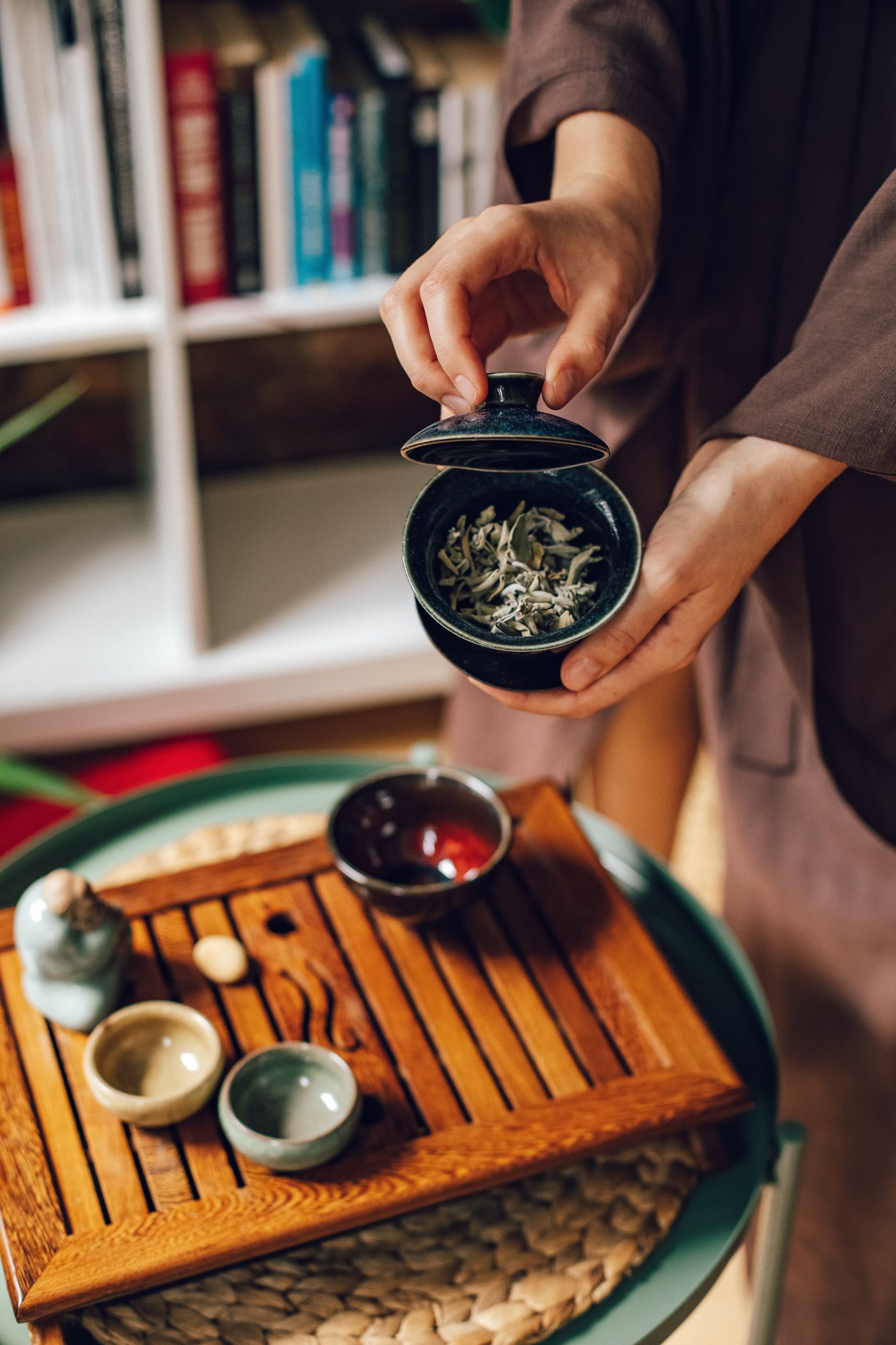 A hand holding a gaiwan above green tea leaves and small tea bowls.
