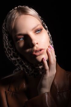 Trendy female in headscarf with leopard print looking away and touching face on black background of studio