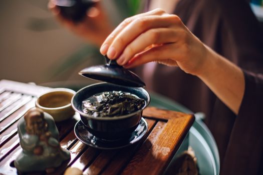 A person's hand lifting the lid of a ceramic bowl during a traditional tea ceremony on a wooden table.
