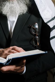 一位老人在室内穿着黑色西装和祈祷披肩，读着一本宗教书籍