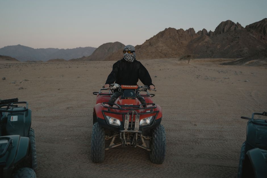 A man wearing a scarf and sunglasses riding an ATV in a rocky desert setting.