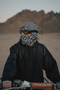 一个戴着护目镜和格子围巾的人，驾驶全地形车穿行于群山环绕的沙漠中，尽显冒险精神