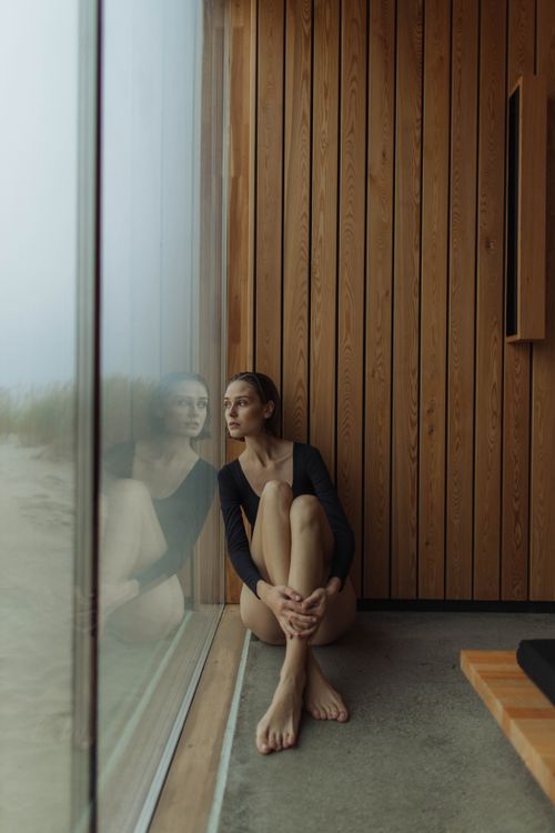 Free Young woman in a black outfit sitting by a large glass window, reflecting deep in thought. Stock Photo