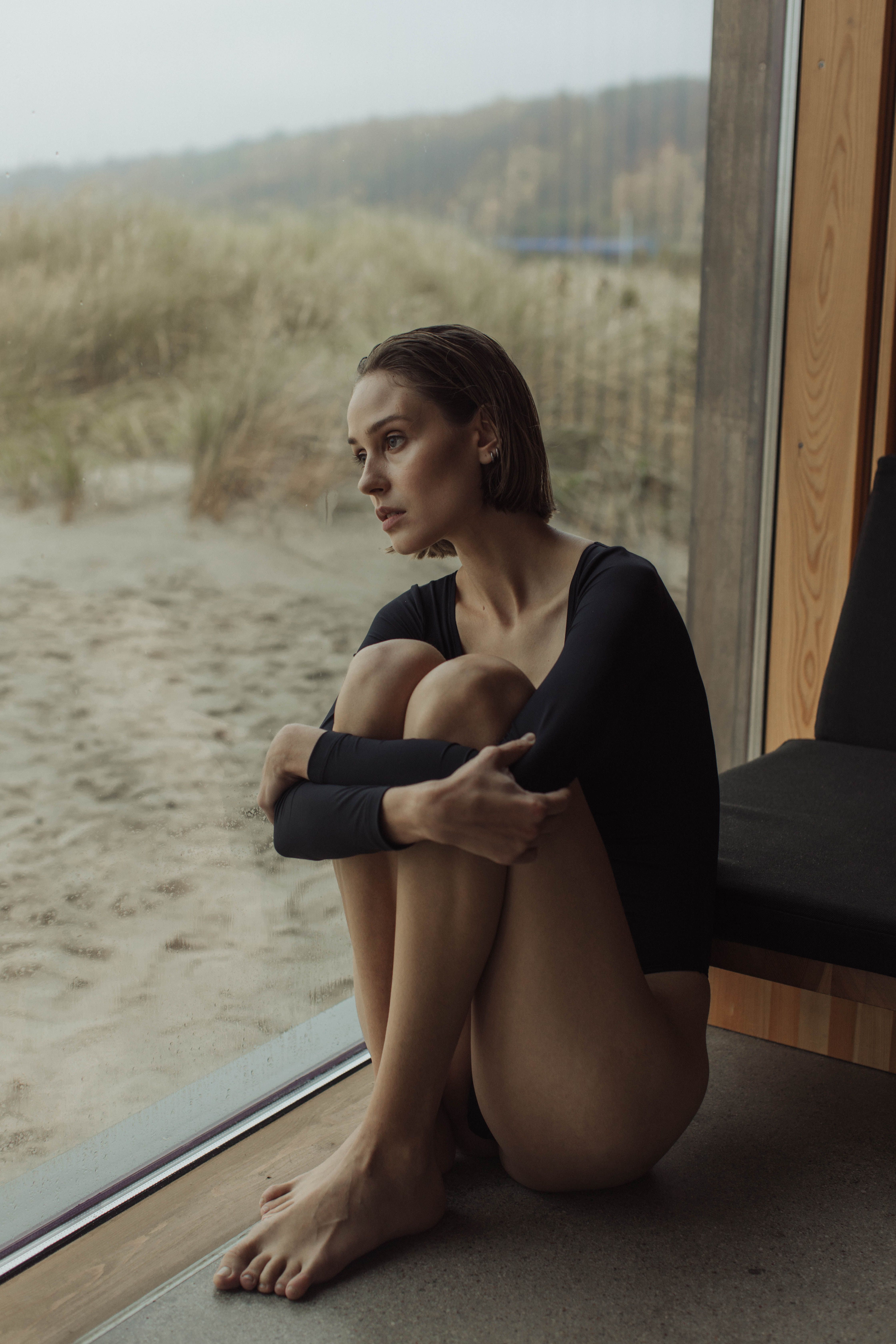 A woman in black long sleeves sits thoughtfully by a glass wall overlooking a sandy landscape.