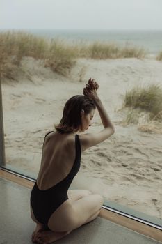 一位身着黑色泳衣的女子，在能望见海滩风景的窗边悠然放松，画面宁静而惬意