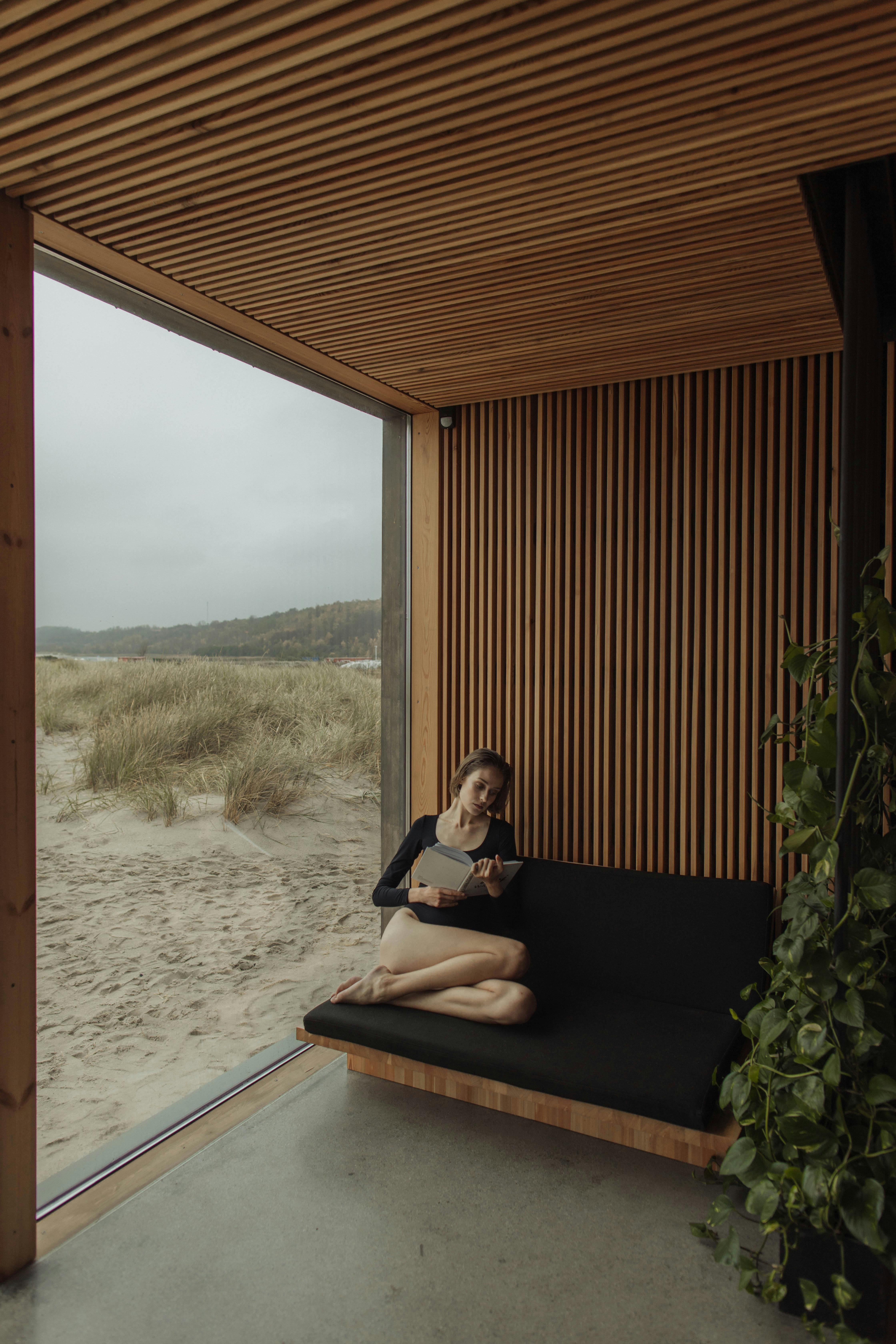 Free Woman reading near a large glass window overlooking a sandy beach. Stock Photo