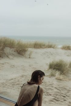 A woman in a backless dress gazes at the serene ocean from a sandy beach.