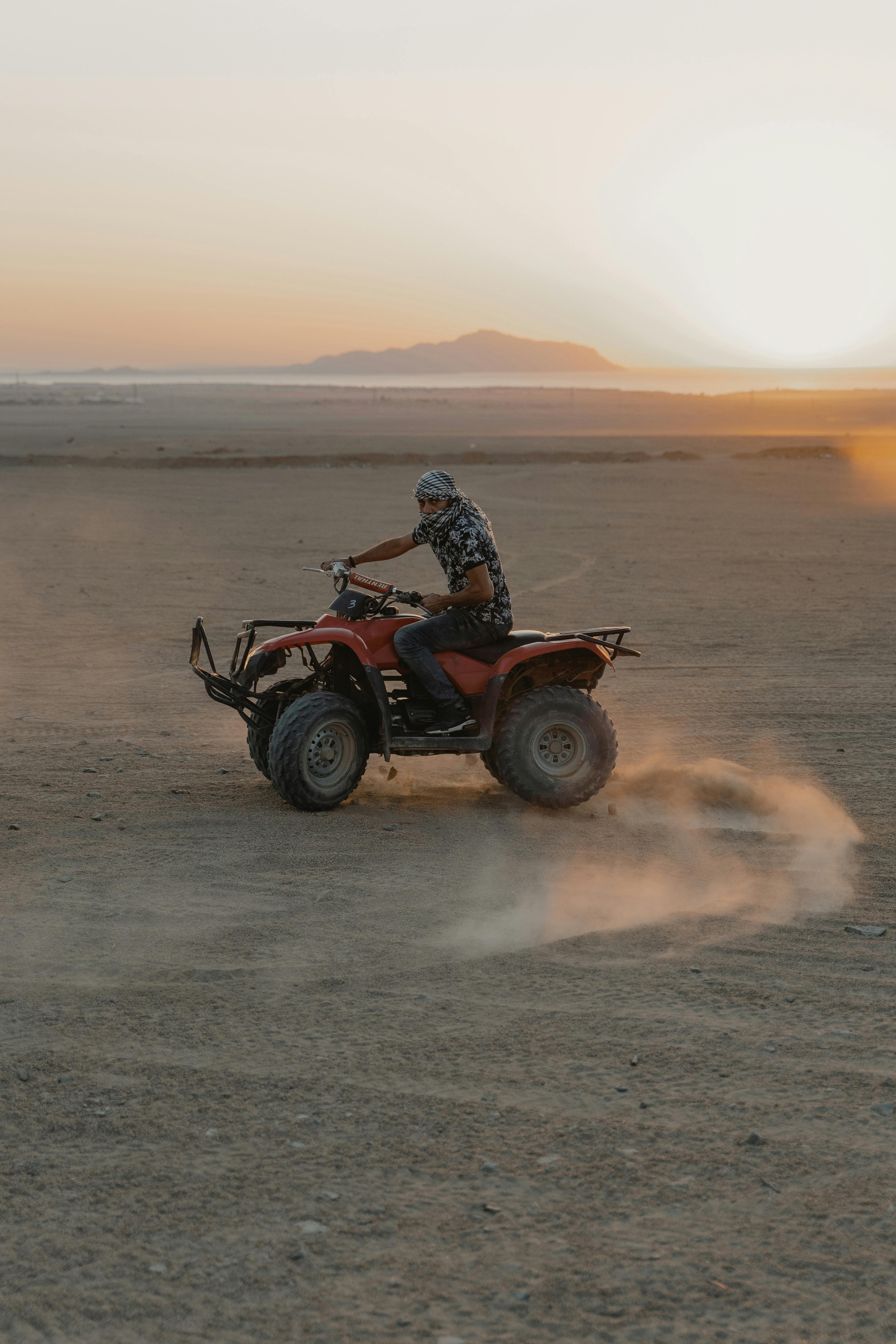 Man riding an ATV across a sandy desert landscape at sunset, kicking up dust.