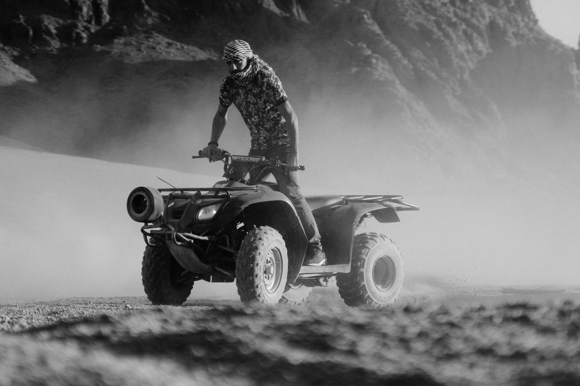 Man riding ATV through a dusty desert landscape, captured in dramatic black and white.