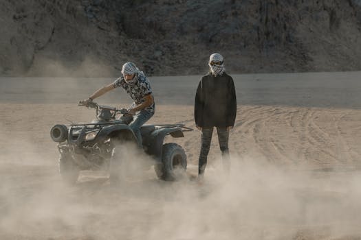 Two individuals in headscarfs riding an ATV through a dusty desert landscape.