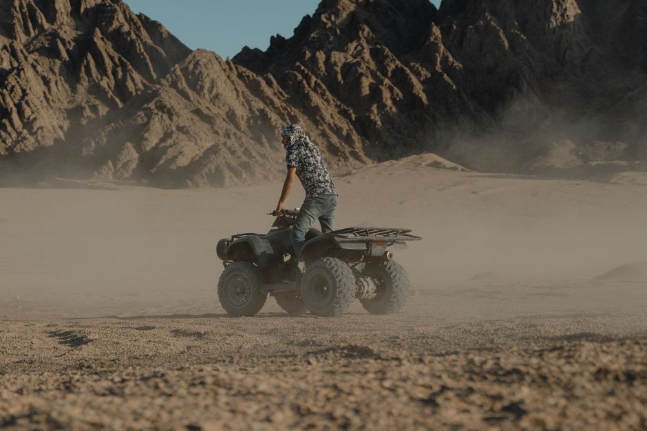 A person riding an ATV across a rugged desert terrain with mountains in the background.