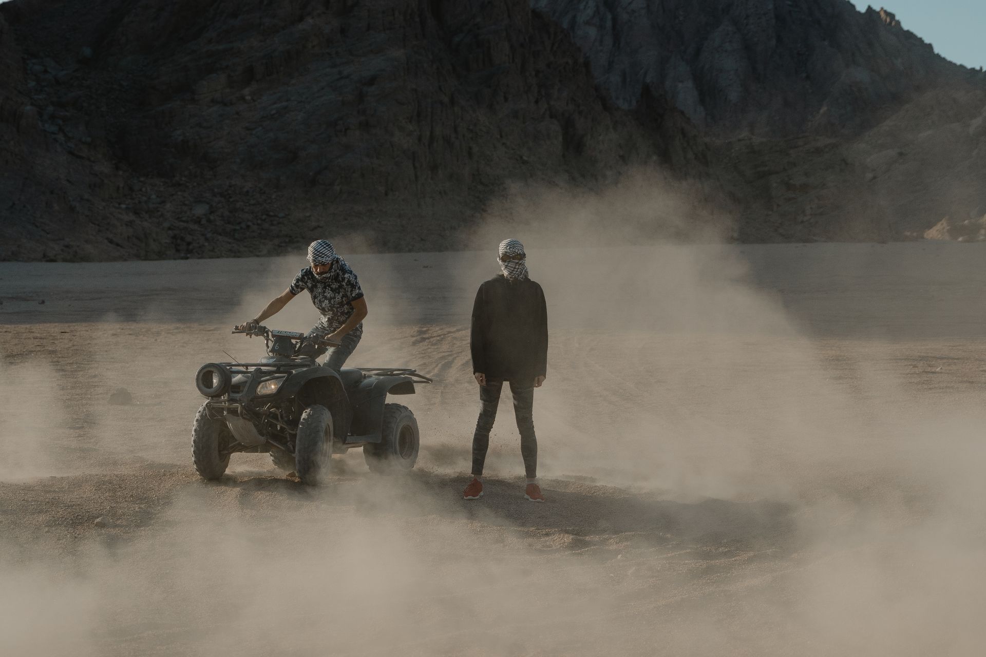 Two people on an ATV stir up dust in a rugged desert landscape, creating a mysterious atmosphere.