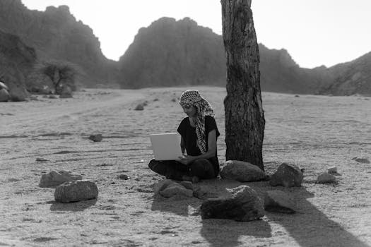 A woman working remotely on a laptop under a tree in a desert landscape.