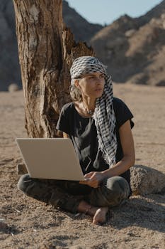 Caucasian woman using a laptop under a tree in a desert setting, wearing a headscarf.