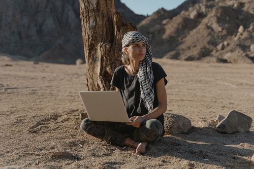 Free Caucasian woman sitting barefoot with laptop under tree in arid landscape. Stock Photo