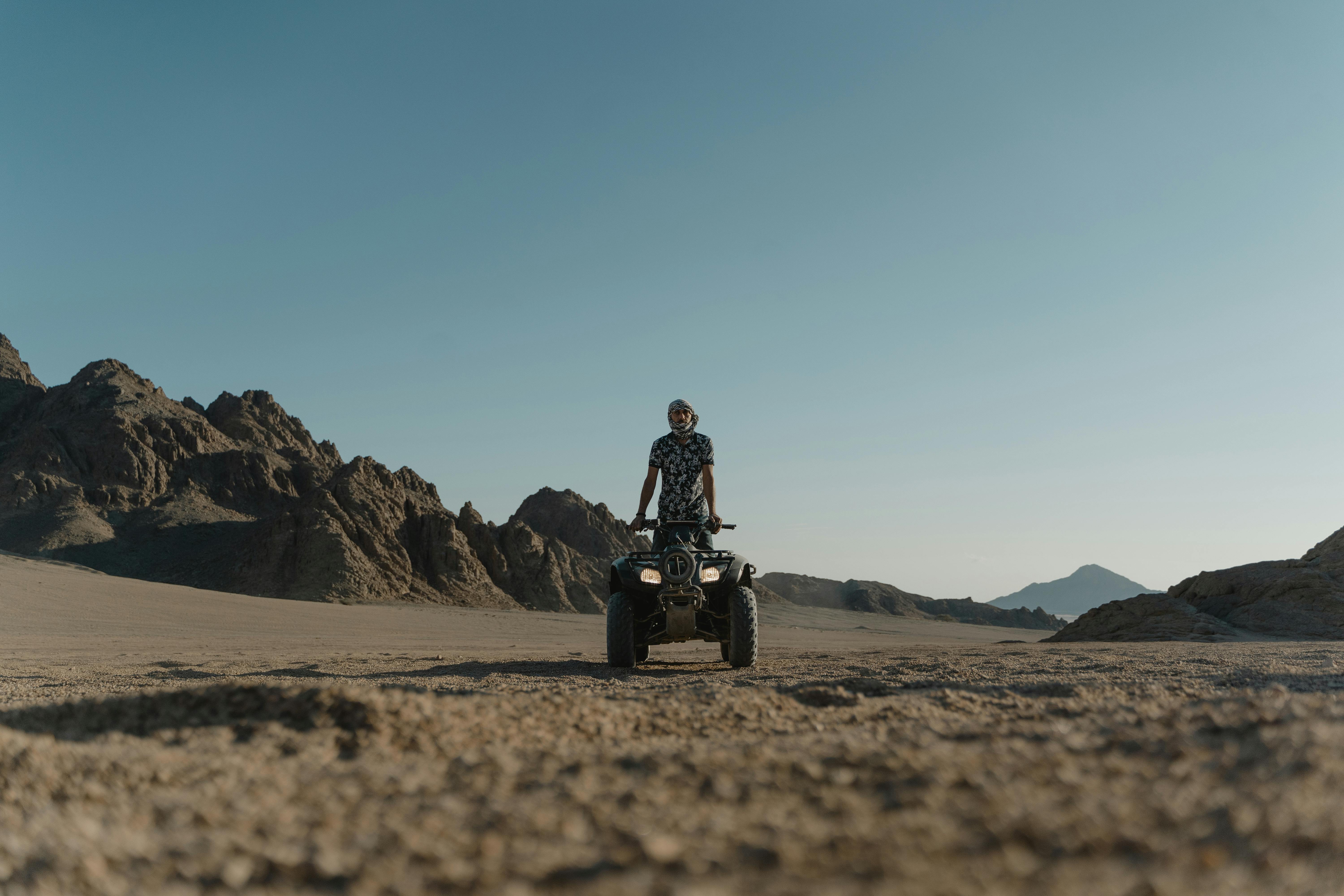 Person standing on a quad bike in a sandy desert with rocky mountains in the background.