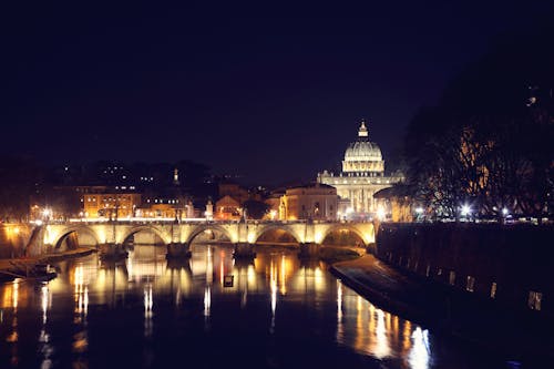 Picturesque night cityscape with dome of famous St Peters Basilica dominating skyline of Rome with illuminate arched bridge over Tiber river