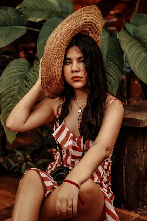 Free Attractive young ethnic female wearing striped summer dress and trendy straw hat sitting in tropical garden and looking at camera Stock Photo