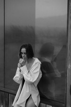 Caucasian woman leaning against a glass wall, lighting a cigarette, in grayscale.
