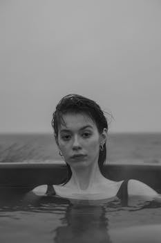 A grayscale portrait of a woman with short hair relaxing in a bathtub overlooking the ocean.