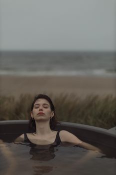 一个女人闭着眼睛，在宁静的海滩背景下的室外按摩浴缸中放松