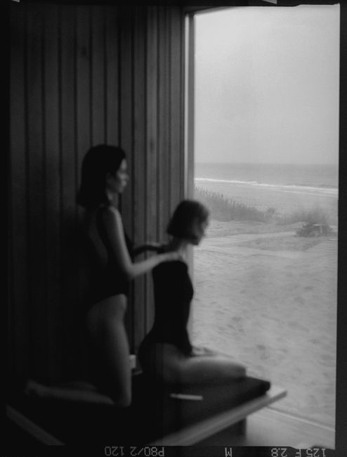 Silhouetted women in swimsuits gazing out at a tranquil beach through a window.