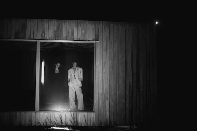 Grayscale Photo Of Two Women Standing By The Glass Window