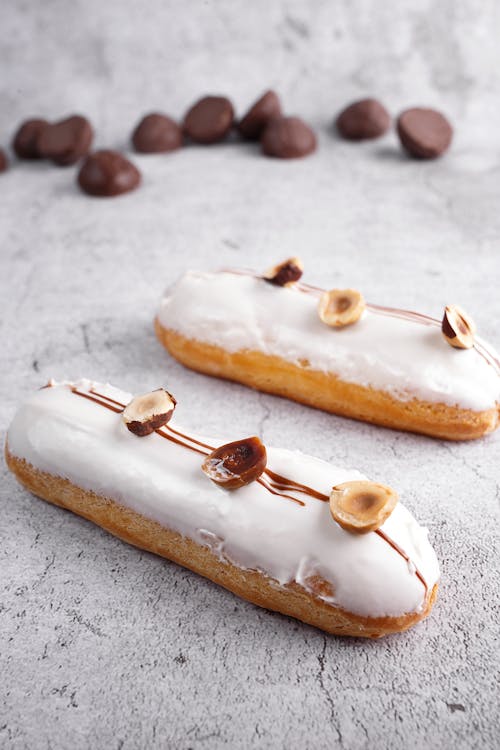 Sweet fresh eclairs topped with whipped vanilla cream and hazelnuts placed on marble tabletop near chocolate chips