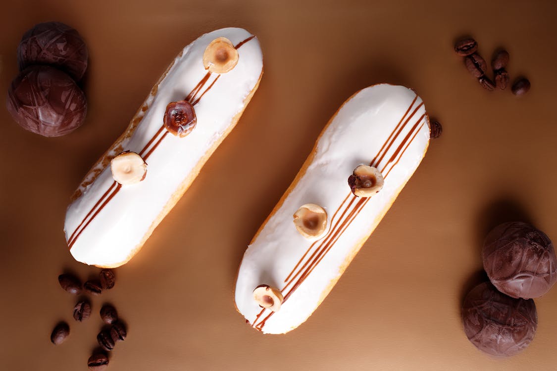 Top view composition of sweet vanilla eclairs topped with hazelnuts and placed on brown table amidst chocolate marshmallow and coffee beans