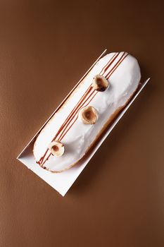 Top view tasty sweet eclair topped with icing and hazelnuts on paper on brown table