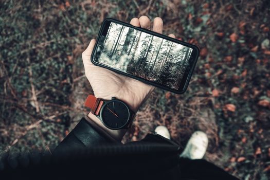 A person outdoors holding a smartphone showing a forest scene, wearing a wristwatch.