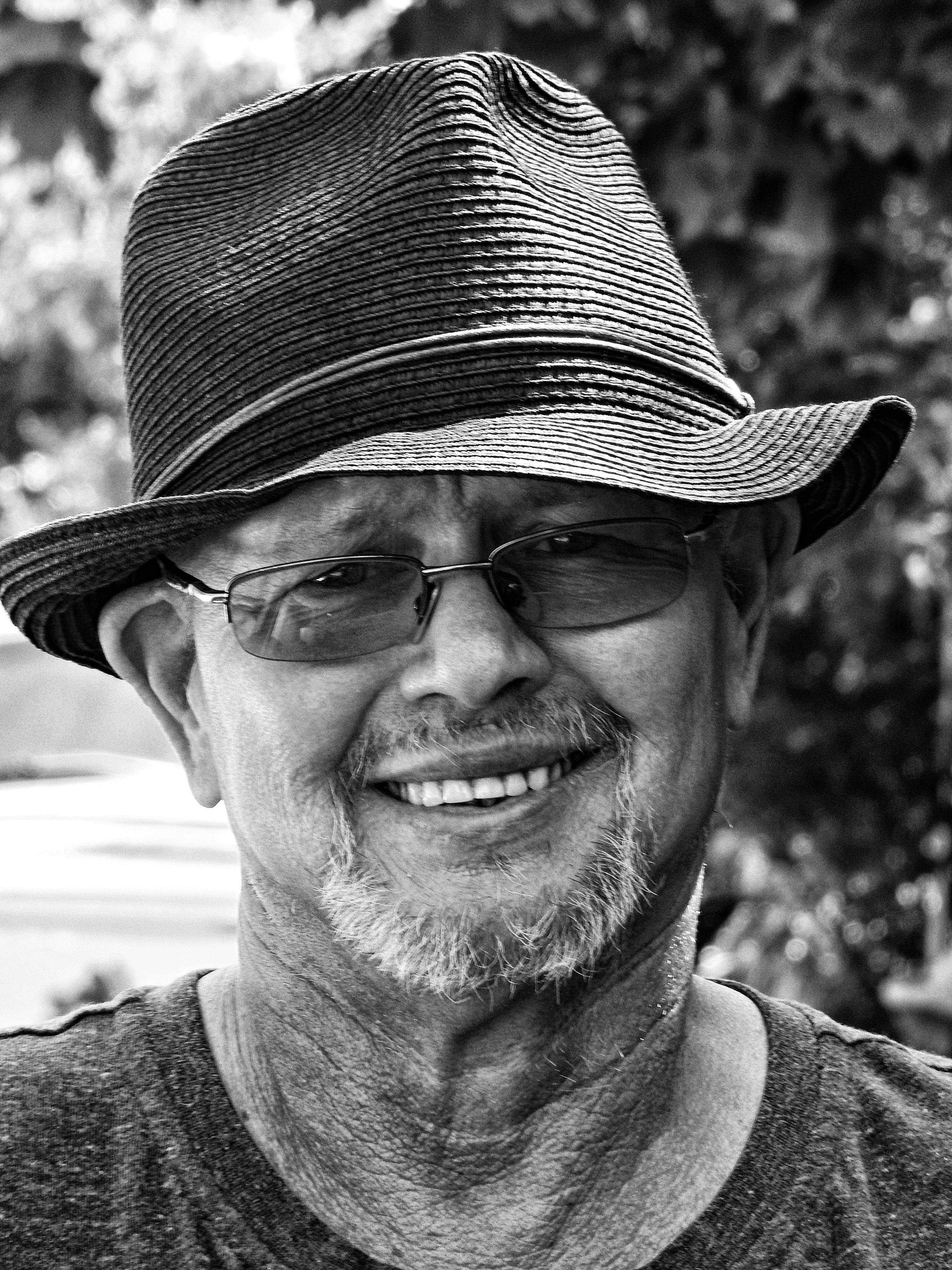 Black and white portrait of an elderly man wearing a hat and glasses, smiling outdoors.