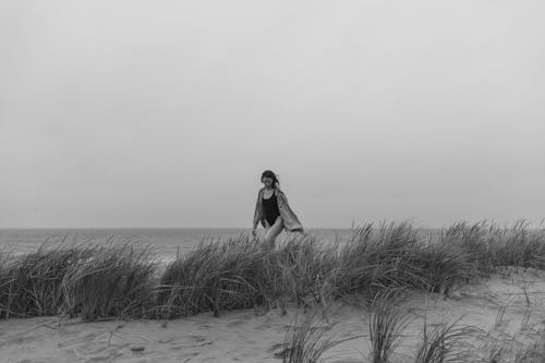 Grayscale Photo of Woman Walking on Beach Sand with Grass 