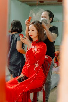 Asian woman in a red traditional dress getting her hair styled indoors, holding a compact mirror.