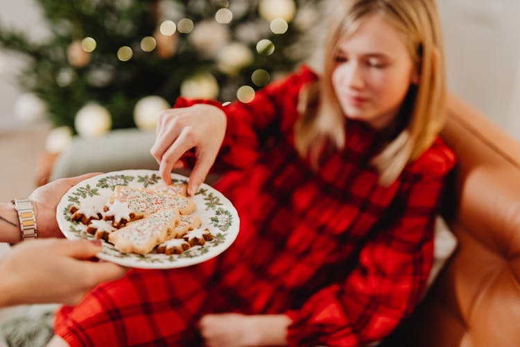 Girl In A Red Checked Dress Taking Ginger Breads From A Plate And Christmas Tree In Background