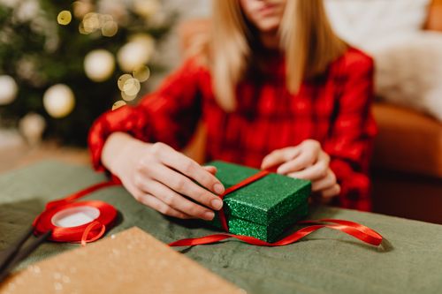 Free Woman wrapping a green glittery gift with red ribbon by a Christmas tree indoors. Stock Photo