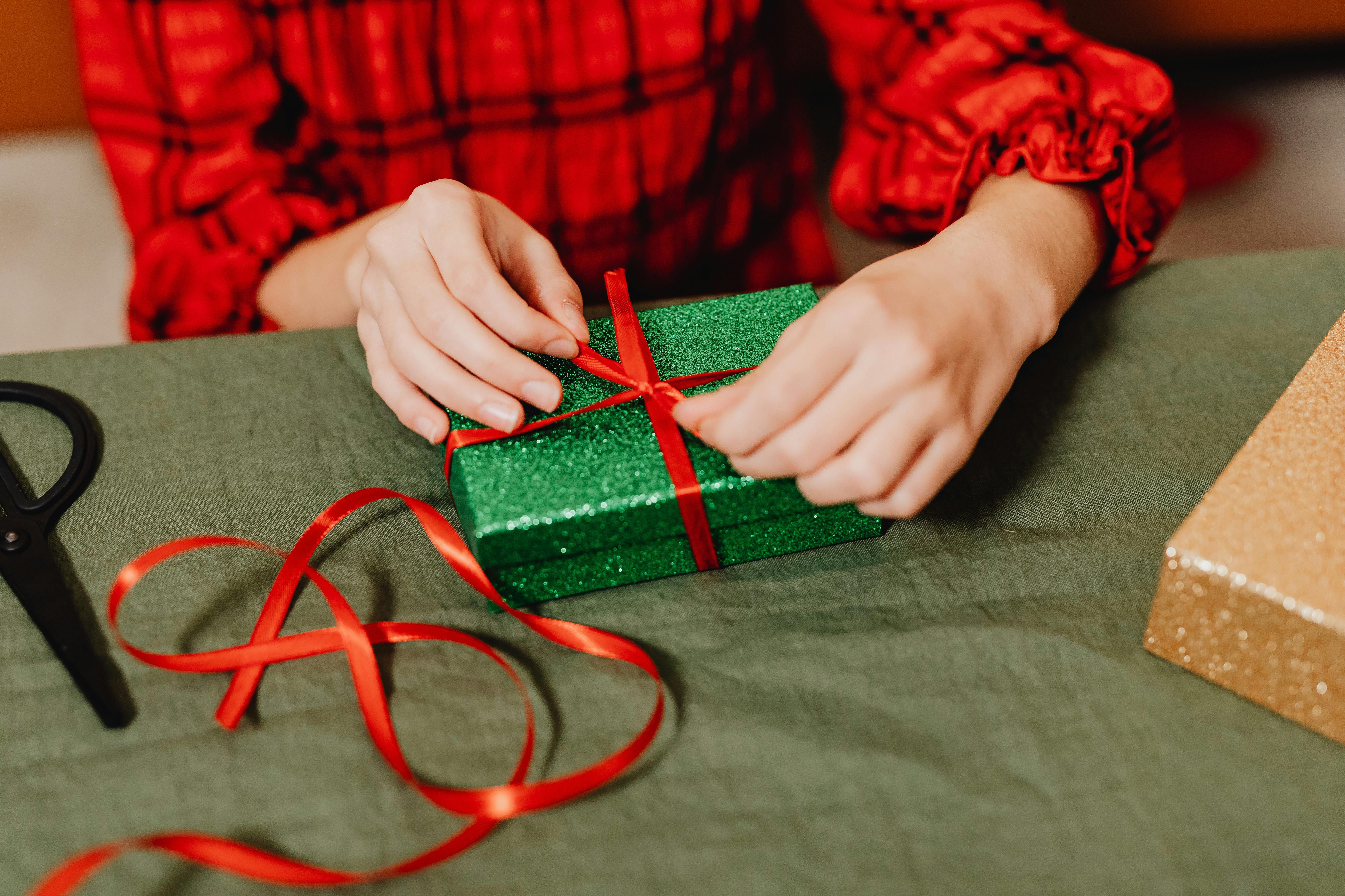 Hands tying a red ribbon on a sparkling green gift box, adding Christmas cheer.