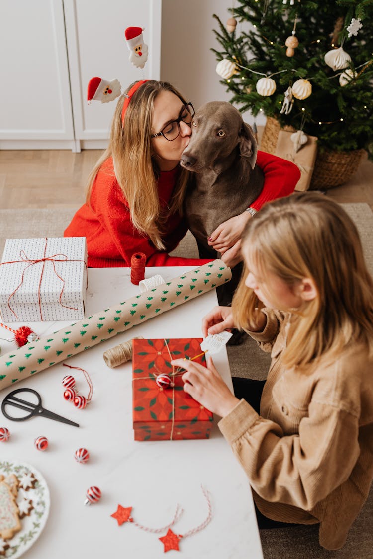 Girl Wrapping Christmas Presents And Woman Kissing A Dog Sitting At A Table